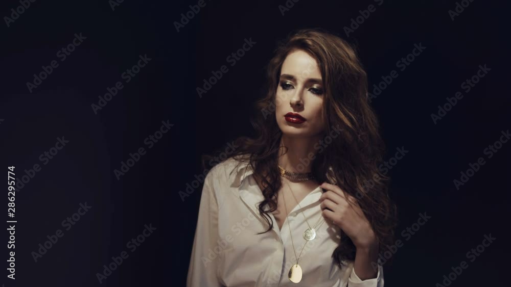 Fashionable beautiful girl in white shirt and with gold jewelry posing on camera. Girl admires her outfit in mirror. Red lipstick. Evening make-up. Hard light and shadow on the girl’s face. Fashionable beautiful girl in white shirt and with gold jewelry posing on camera. Girl admires her outfit in mirror. Red lipstick. Evening make-up. Hard light and shadow on the girl’s face.