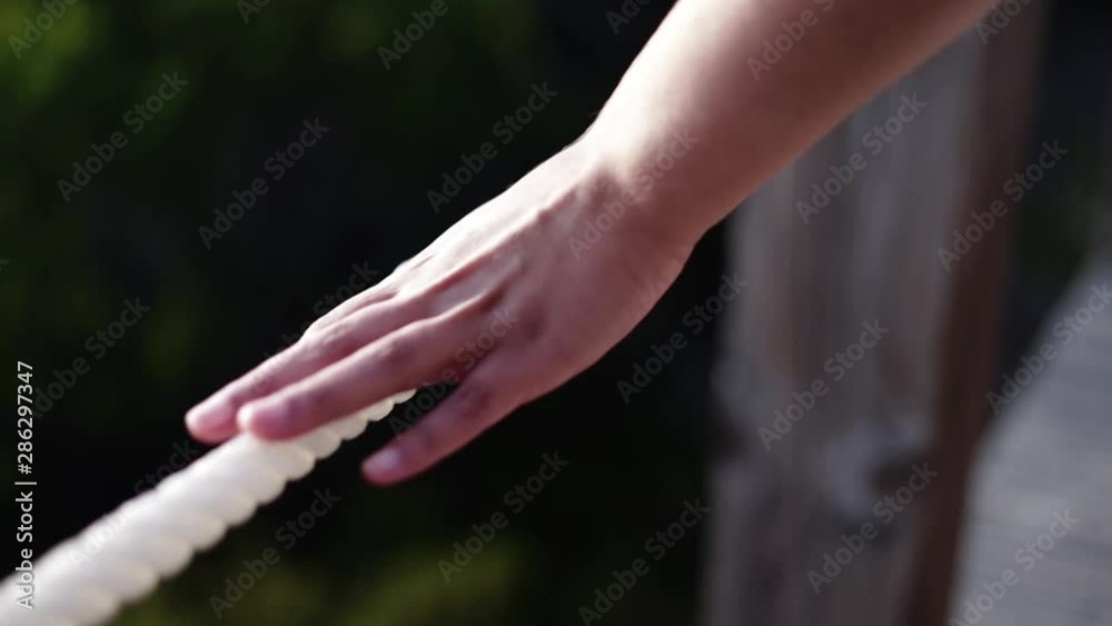 Acompanhamento de mão de mulher a andar e a tocar numa corda branca de um passadiço de madeira com uma lente de grande desfoco.