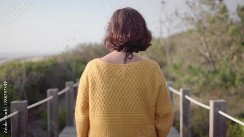 Acompanhamento de mulher com casaco amarelo vista de costas a andar pelo passadiço de madeira com muita natureza envolvente. Em Portugal junto à praia. 