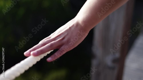 Acompanhamento de mão de mulher a andar e a tocar numa corda branca de um passadiço de madeira com uma lente de grande desfoco.