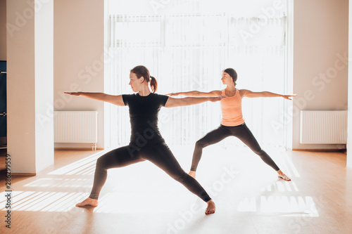 Two attractive young women balancing and practicing yoga in a light studio. Well being, wellness concept. One of the woman is plus size. Varrior pose