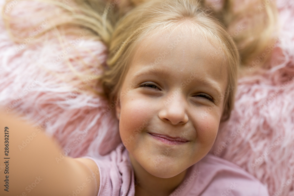 Adorable little girl with long blonde hair lying on pink fur blanket and making selfie. Positive emotions , expression, fun. Happy smiling kid, childhood day, education, kids using technology concept