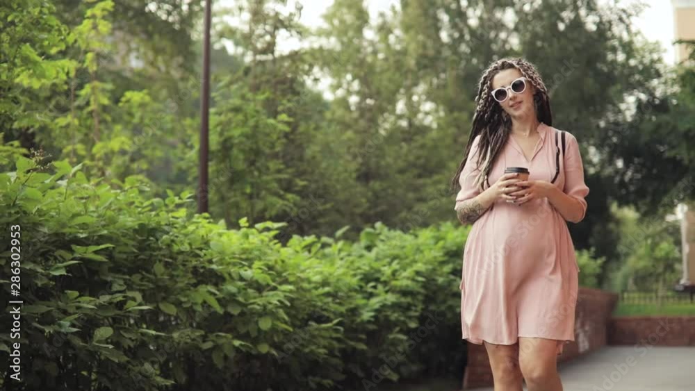 Maternity leave. Pregnant happy young woman hipster walking in city park with coffee in hands. Woman enjoying her pregnancy expecting a baby at summer warm day. She is in cute dress with braids.