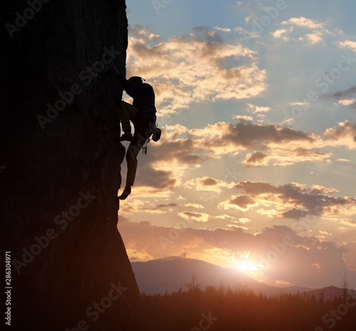 Side view of strong rock climber at dawn. Male silhouette on high rocky wall having extreme outdoors activity. Concept of perseverance and never give up. Sunrise sky with clouds and copy space