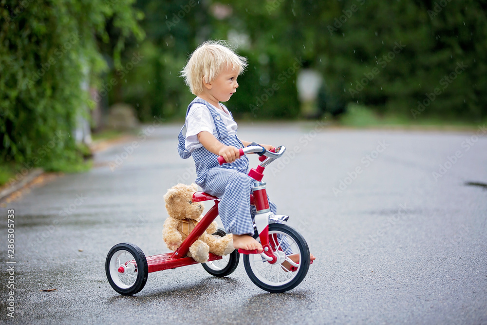 Cute little boy, with teddy bear toy, riding tricycle on the street in the rain, barefeet Cute little boy, with teddy bear toy, riding tricycle on the street in the rain, barefeet