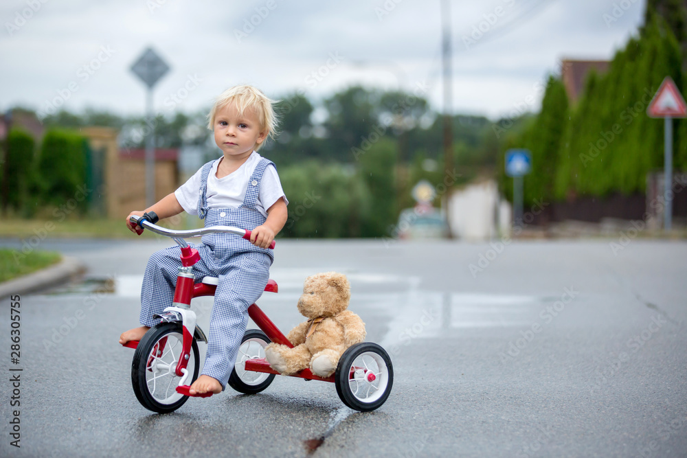 Cute little boy, with teddy bear toy, riding tricycle on the street in the rain, barefeet Cute little boy, with teddy bear toy, riding tricycle on the street in the rain, barefeet
