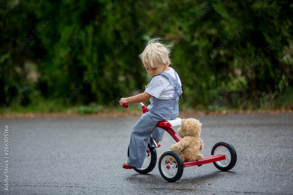 Cute little boy, with teddy bear toy, riding tricycle on the street in the rain, barefeet Cute little boy, with teddy bear toy, riding tricycle on the street in the rain, barefeet