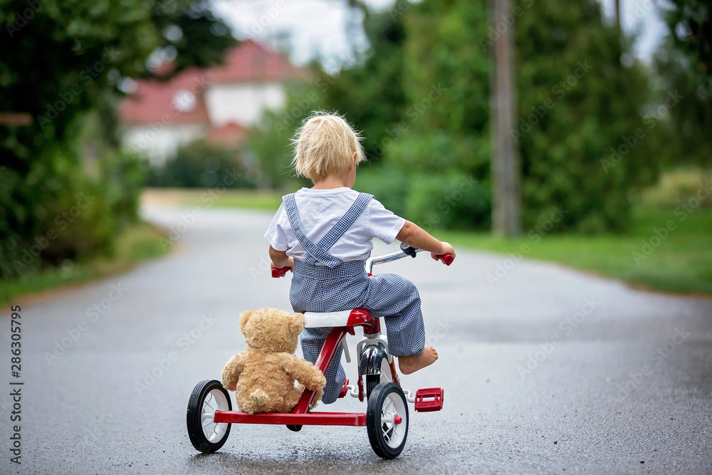 Cute little boy, with teddy bear toy, riding tricycle on the street in the rain, barefeet Cute little boy, with teddy bear toy, riding tricycle on the street in the rain, barefeet