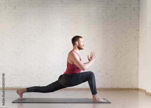 Warrior pose. Caucasian fit man stands in deep stretching and doing yoga in fitness studio, side view, selective focus.