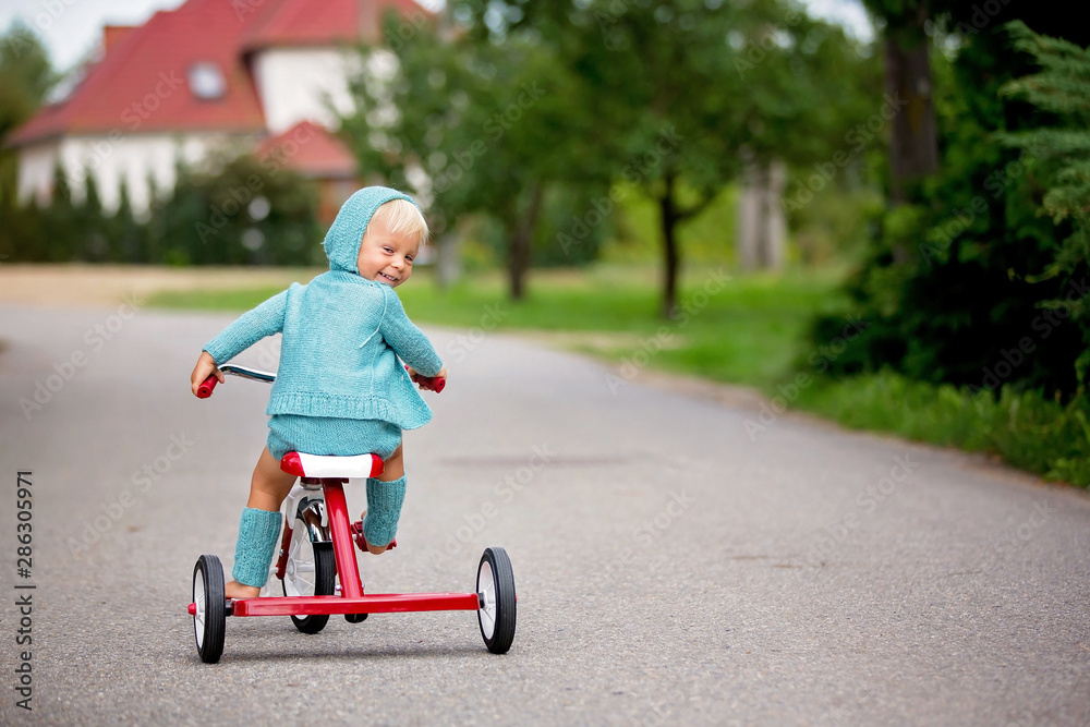 Adorable toddler boy with knitted outfit, riding tricycle on a quiet village street Adorable toddler boy with knitted outfit, riding tricycle on a quiet village street