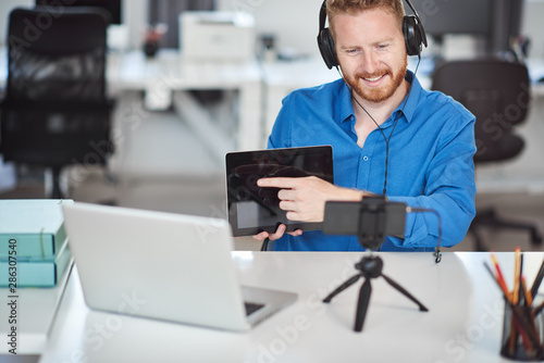 Young Caucasian hardworking employee havining video call over smart phone. In hands is tablet and on head are headphones. Remote business concept.