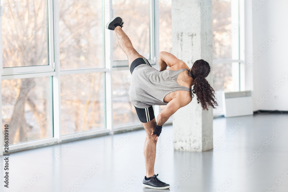 Fototapeta premium Back side of healthy muscular young adult man with curly long hair is posing on professional kickboxing position with raised legs, showing combat technique. Indoor, window background, martial arts