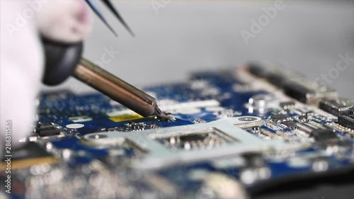 Technician engineer in workshop. Repairman in gloves is soldering circuit board of electronic device on the table, close up. He takes tin with a soldering iron and puts it on microcircuit, slow motion