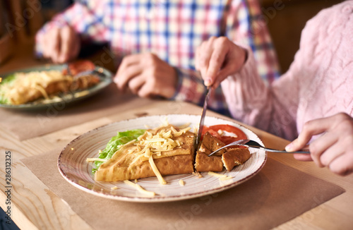Focus on dish served with vegetables and cheese. Delicious crepe sandwich lying on wooden table near cropped female next to male on blurred background. Woman with fork and knife during lunch in cafe. © anatoliy_gleb