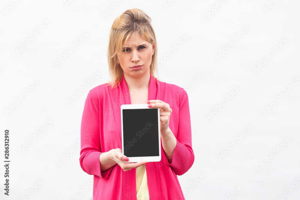 Portrait of dissatisfied beautiful young businesswoman in pink blouse standing and holding tablet empty screen and looking at camera with unhappy face. Indoor, isolated, studio shot, white background