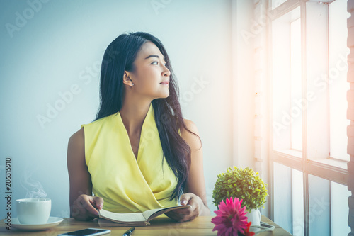 Asian girl with long hair drinking coffee and reading pocket book by the window. She smiled at the mouth with eyes looking outward in the solar atmosphere evening. Concept of education and learning.