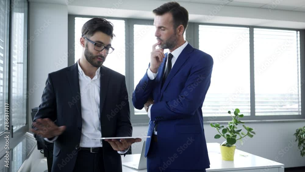 Two businessmen standing in office with tablet discussing business strategies and then shaking hands Two businessmen standing in office with tablet discussing business strategies and then shaking hands