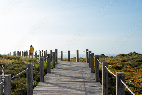 Woman from the back with a yellow jacket in a distance standing on a wooden walkway, contemplating the blue sky
