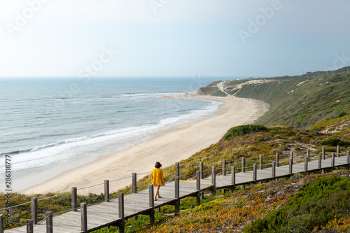 Girl in a distance walking in a long wooden walkway looking at the beach below. Surrounded by vegetation and the sea.
