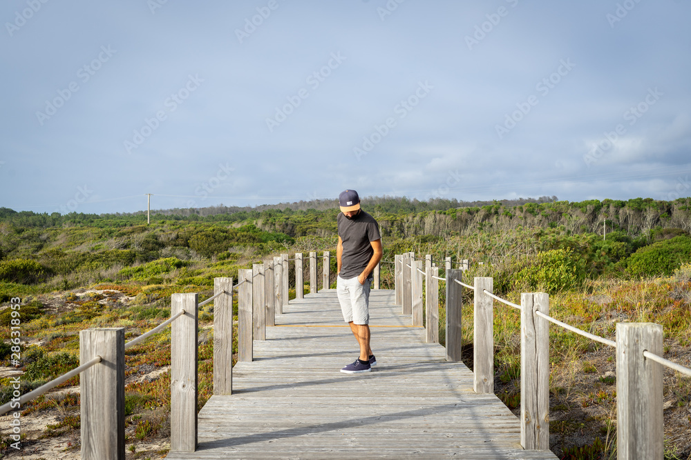 Bearded Man wearing a cap view from the front, looking down, in a distance standing on a wooden walkway surrounded by nature and a forest on the background.