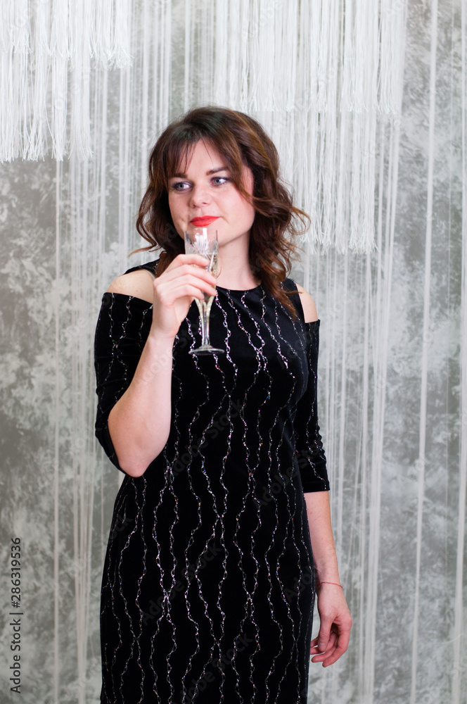Obraz premium Very beautiful brown-haired woman with curly hair in an evening dress with a glass of champagne against the gray wall