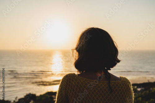 Girl with short hair and a yellow sweater watching the sunset on the beach below.