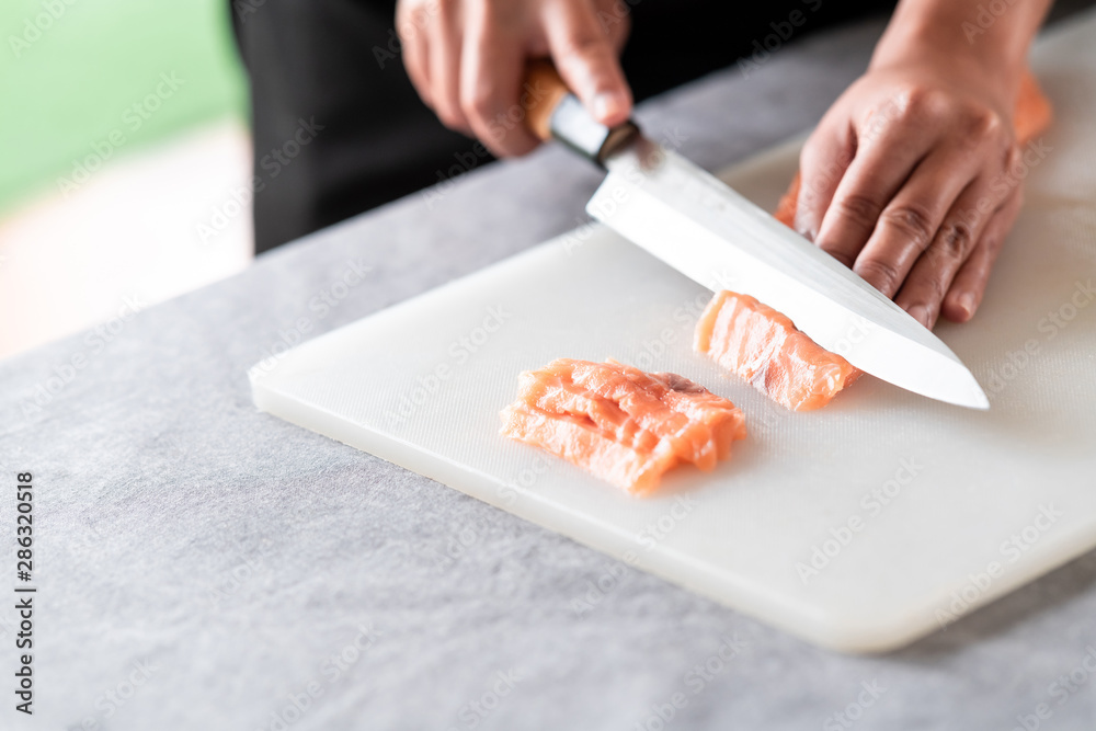 Chef slicing raw salmon on plastic plate. Asian woman chef in black uniform, slicing small piece of salmon. Complete pieces on side with green floor.