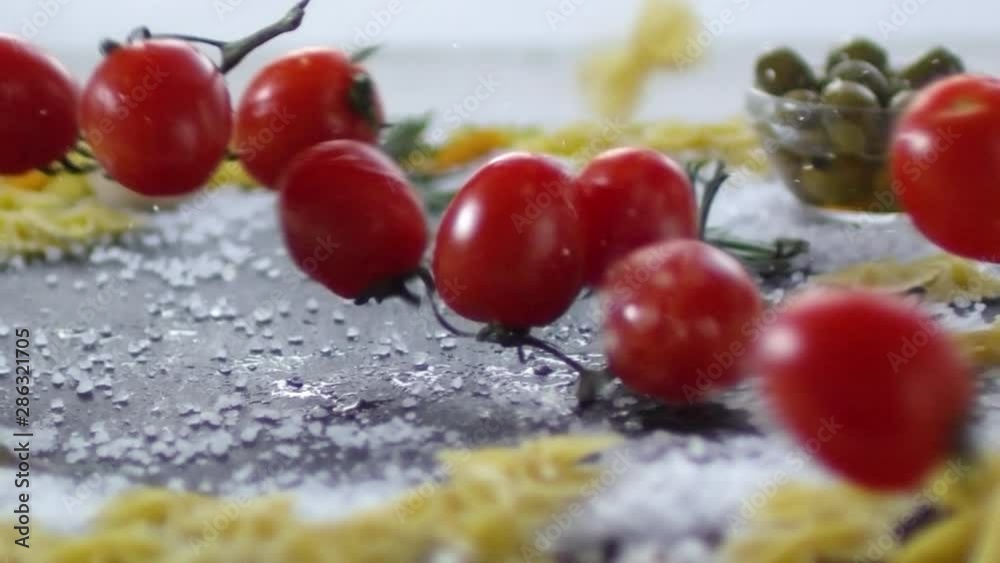 Close up tracking shot of fresh cherry tomatoes falling down with water droplets on kitchen table with pasta, salt, olives and herbs on it Close up tracking shot of fresh cherry tomatoes falling down with water droplets on kitchen table with pasta, salt, olives and herbs on it