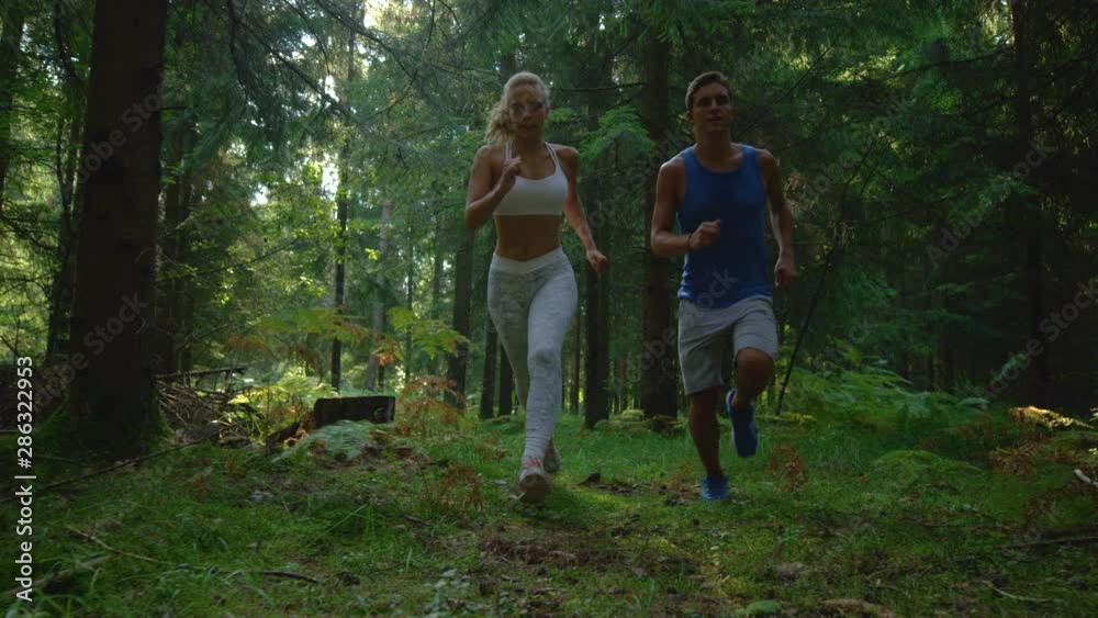 CLOSE UP, SLOW MOTION, LOW ANGLE: Active Caucasian man takes his girlfriend for a scenic jog in the sunny woods. Carefree sporty couple jogs along the sunlit forest trail on a perfect summer day.