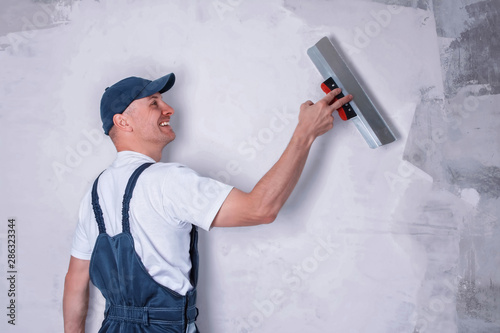 Fotografía Smiling worker in profile wearing overalls and cap plastering a wall with finishing putty using a putty knife