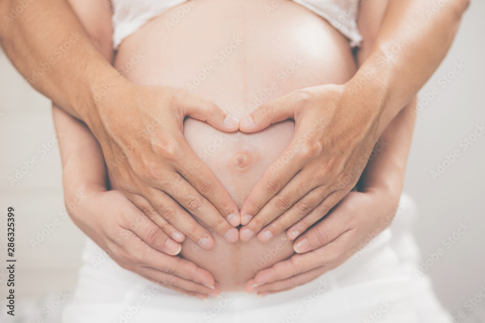 pregnant woman and her husband hand splice hugging the tummy Heart shaped show love for the unborn baby in the belly relaxing at home in bedroom. wait birth date expecting a baby stand in the outdoors