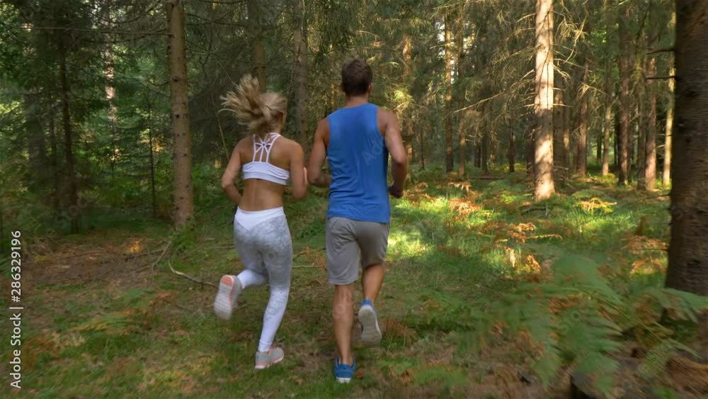 Unrecognizable blonde haired woman in white sportswear and her athletic boyfriend jog through the peaceful pine forest. Young sporty couple training together in the woods for a trail running marathon. Unrecognizable blonde haired woman in white sportswear and her athletic boyfriend jog through the peaceful pine forest. Young sporty couple training together in the woods for a trail running marathon.