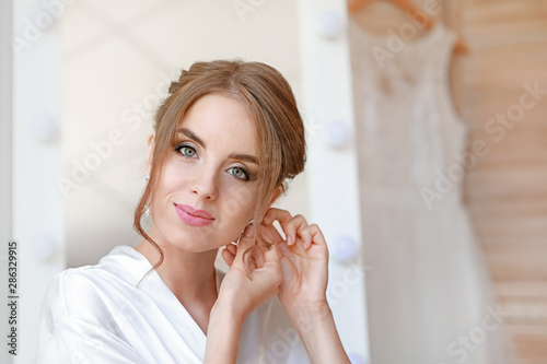 Beautiful young bride wearing earrings before wedding ceremony at home