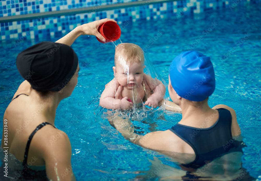 Front view of baby boy during swimming lesson. Back view of mother holding child and woman pouring water on child's head, accustoming his to water. Formation of hardiness and endurance in infants