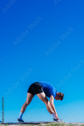 Blonde athlete wearing blue dry-fit shirt stretching and preparing for a workout while standing on top of a cliff which has an amazing view.