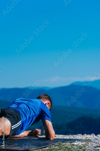 Strong male athlete doing plank while exercising his abs on top of a mountain on a sunny day.
