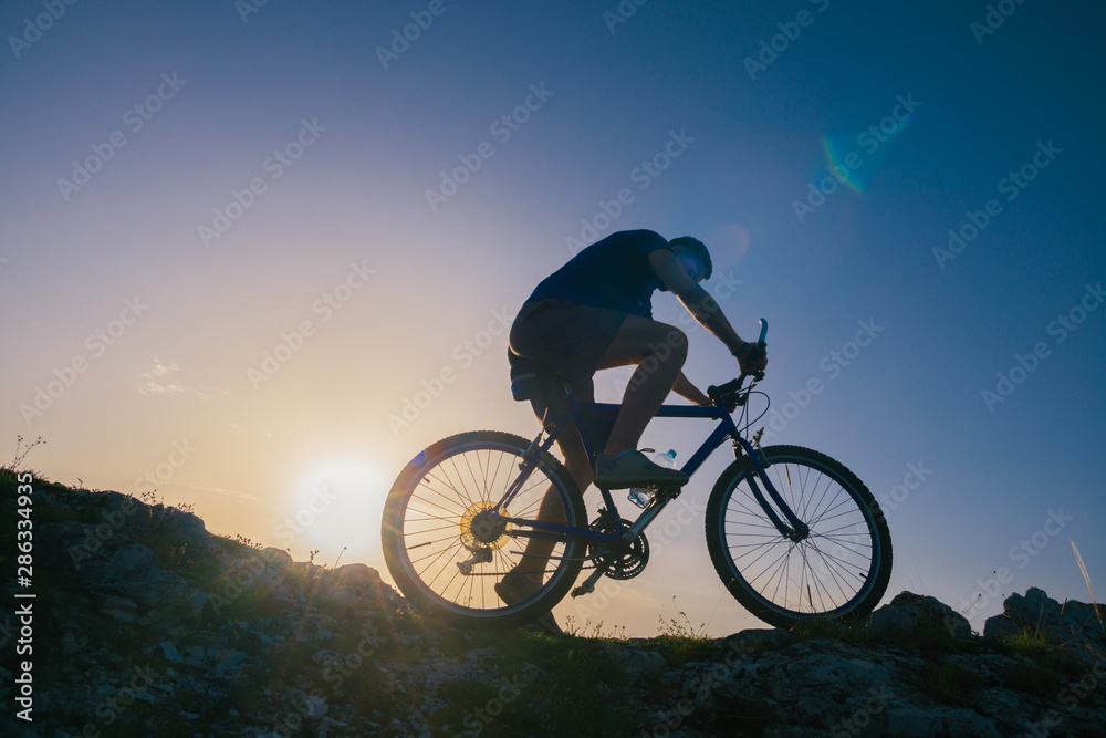 Naklejka premium Silhouette of a fit male mountain biker riding his bike uphill on rocky harsh terrain on a sunset.