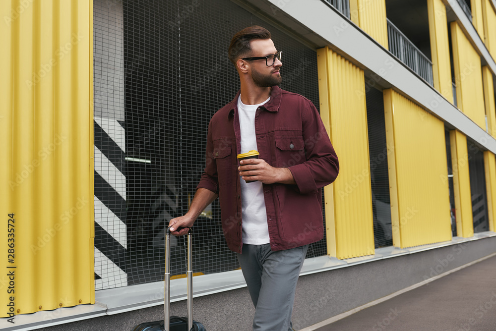 Tourist. Handsome bearded man in casual wear and eyeglasses holding a disposable cup and carrying his luggage while walking through the city street - obrazy, fototapety, plakaty 