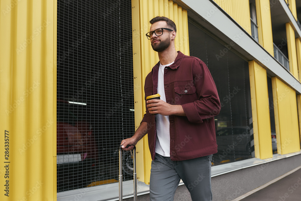 Time to travel. Handsome smiling bearded man in casual wear and eyeglasses holding a disposable cup and carrying his luggage while walking through the city street