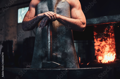 Billede på lærred Muscular blacksmith in protective apron at his workshop with hammer in his hands