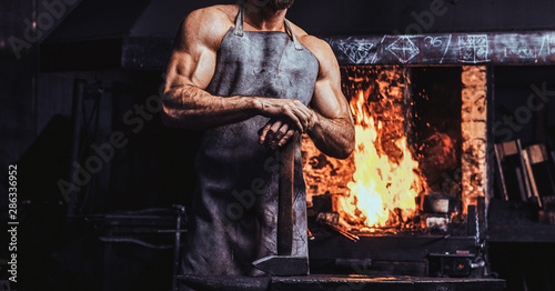 Fotografia Muscular blacksmith in protective apron at his workshop with hammer in his hands