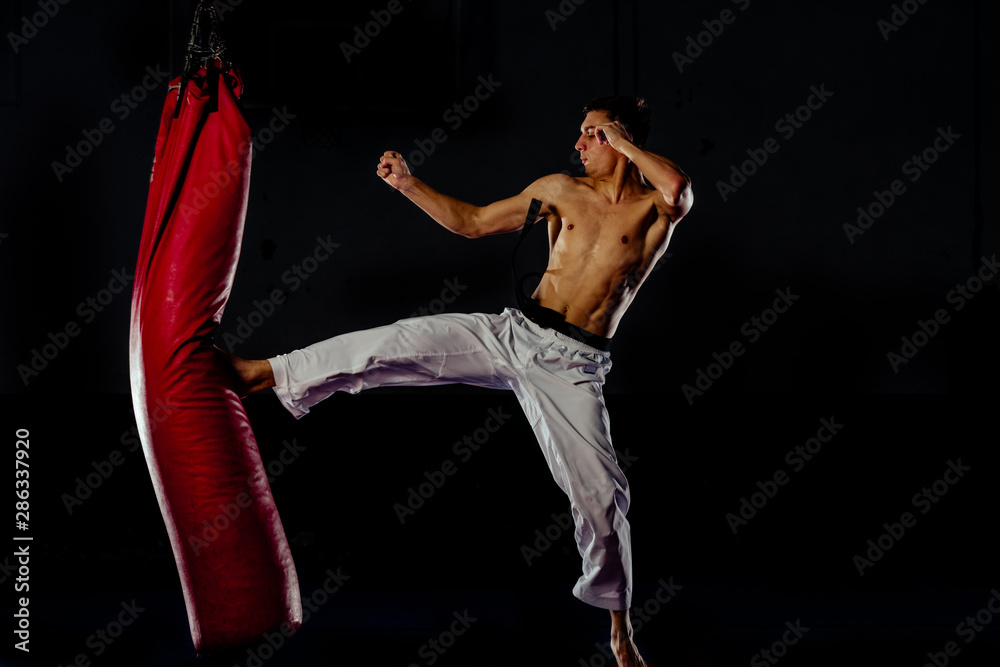 Athletic man kicking punching bag with leg, training kickboxing on the heavy red sand bag
