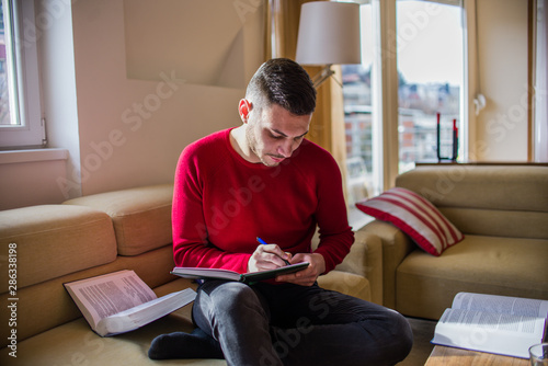 A young man is sitting on sofa and is writing in a notebook