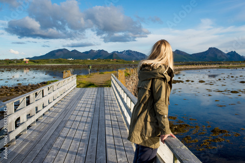 A girl stands on a wooden bridge and looks forward. Beautiful nature landscape in North. Scenic outdoors view. Water, mountains and clouds. Travel, adventure, lifestyle. Explore Norway, summer © Iuliia Pilipeichenko