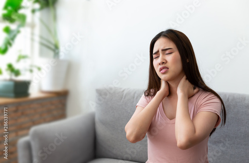 Asian young woman was holding her hand to catch her neck, she has a sore throat sitting on the sofa at home.