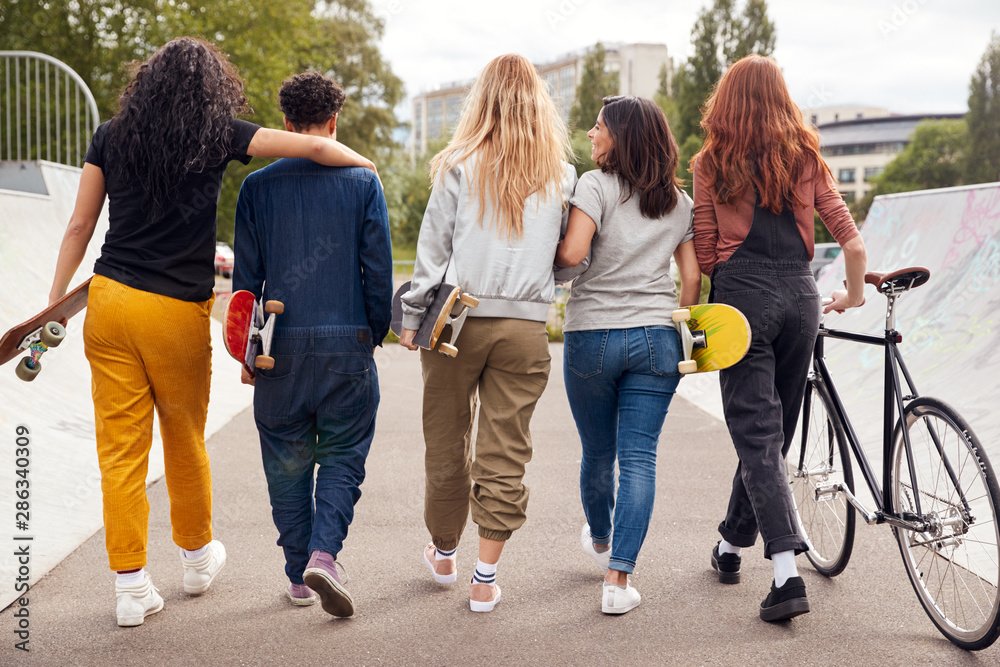 Obraz premium Rear View Of Female Friends With Skateboards And Bike Walking Through Urban Skate Park