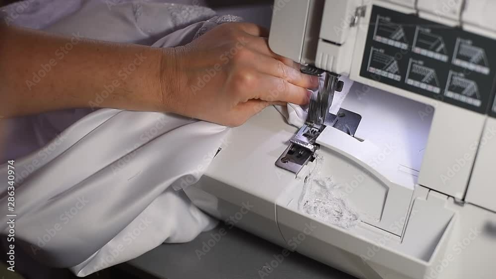 Focus on stitching process on manufacturing backlit sewing machine overlock. Cropped female hands doing seam for overing edge of cloth. Cutting off white thread after finishing working operation. POV