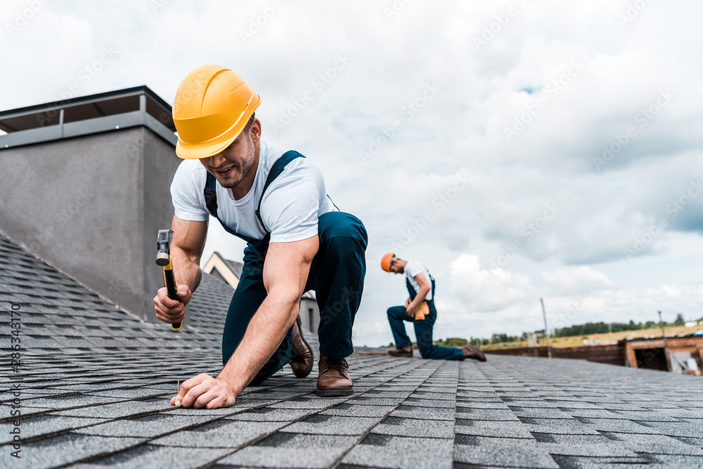 selective focus of handyman holding hammer while repairing roof near coworker - obrazy, fototapety, plakaty 