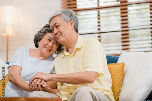 Asian elderly couple holding their hands while taking together in living room, couple feeling happy share and support each other lying on sofa at home. Lifestyle Senior family at home concept.