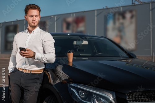 Young successful businessman in a white shirt checking smartphone standing near his luxary car. Disposable cup of hot coffee on a motor hood. © ianachyrva